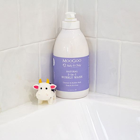 A bottle of MooGoo Baby & Child Natural 2-in-1 Bubbly Wash sits on a white bathtub ledge against a white tiled wall, with a small cow-shaped soap dispenser beside it. Hypoallergenic and SLS free to soothe sensitive skin. 