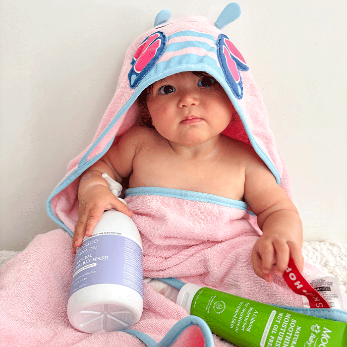 Baby wrapped in a pink hooded towel holding a bottle of MooGoo Bubbly wash and MooGoo Soothing Nut Oil free Moisturiser. 