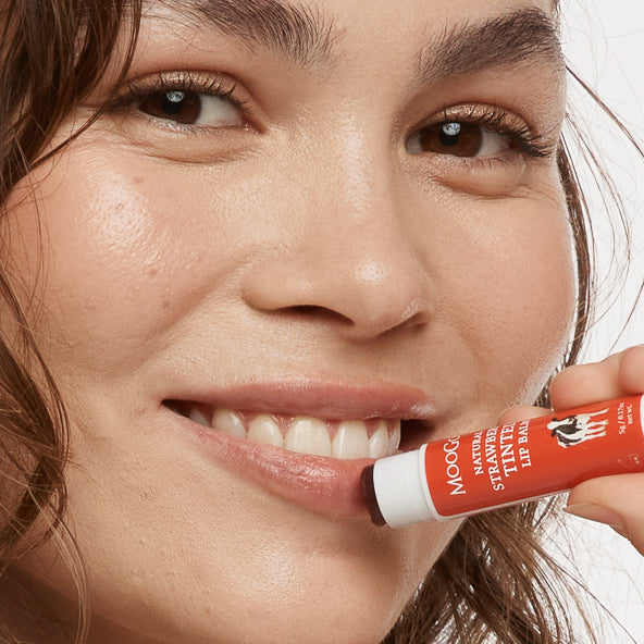 Woman applying MooGoo Strawberry Tinted Lip Balm  white background, natural lip care with edible oils, soft hydration and a gentle strawberry flavour, phthalate free, petroleum free, beetle and synthetic dye free. 