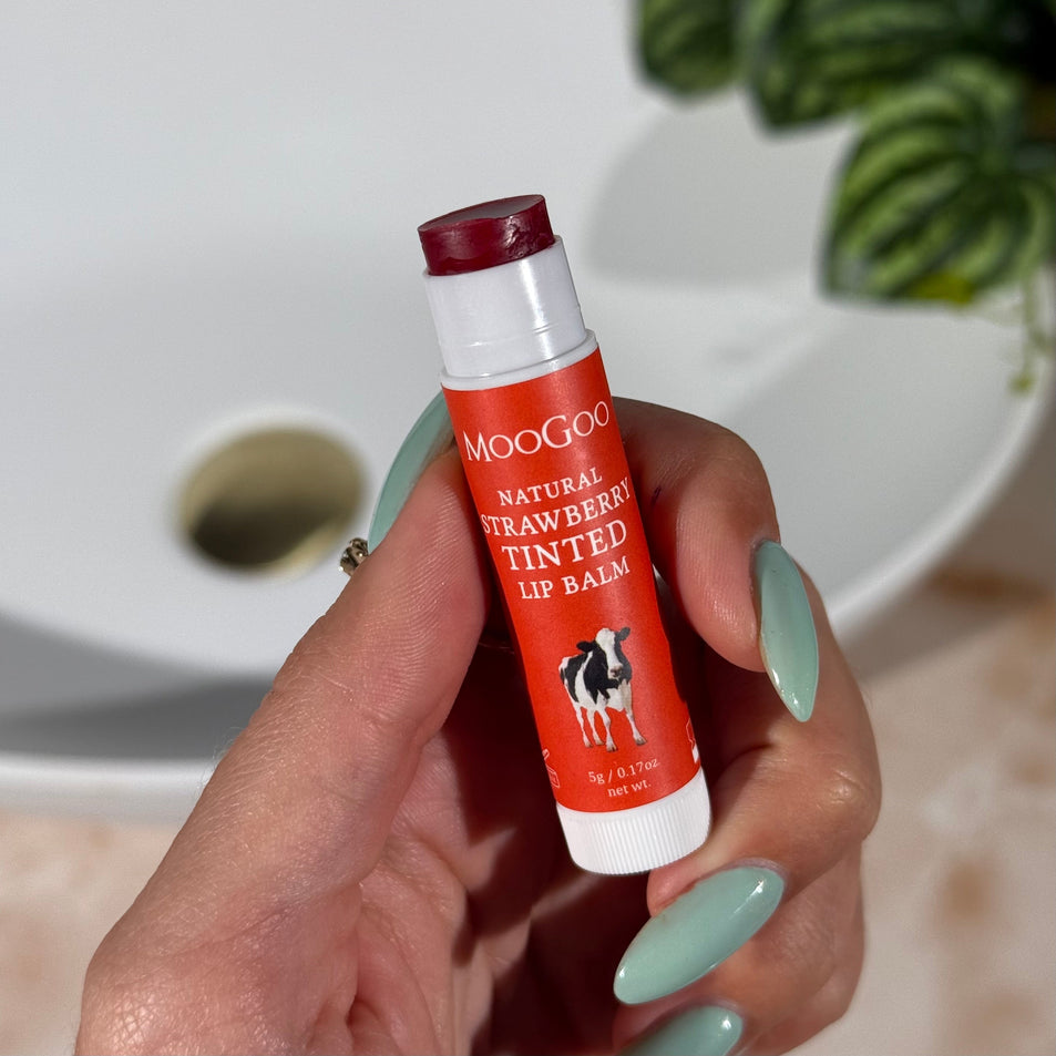 Hand holding a red tube of MooGoo Natural Strawberry Tinted Lip Balm with a plant and bathroom sink in the background.