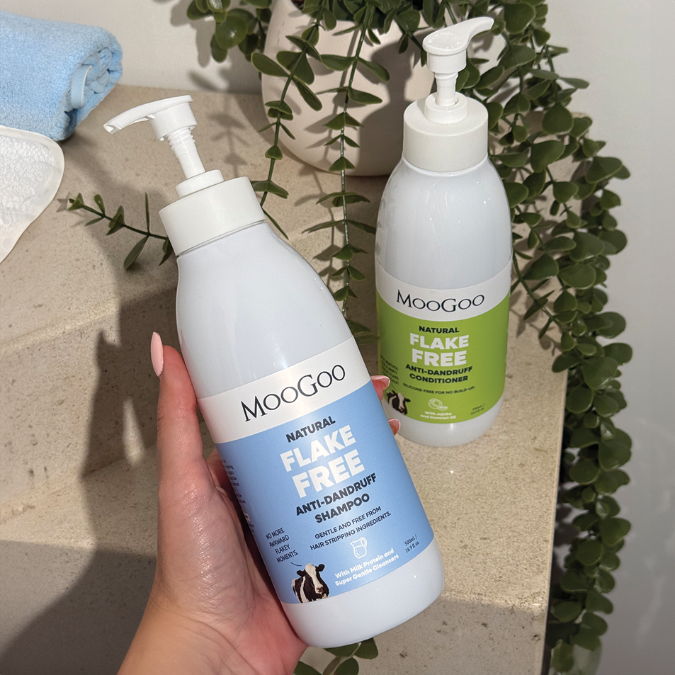a hand holding MooGoo MooGoo Flake Free Anti-Dandruff Shampoo and Conditioner, with towels and greenery in the background, silicone free formula with jojoba and coconut oil to restore dry scalp